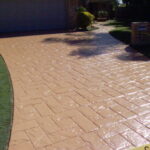 A glossy, tan-coloured stamped concrete driveway with a rectangular brick-like pattern curving toward a house.