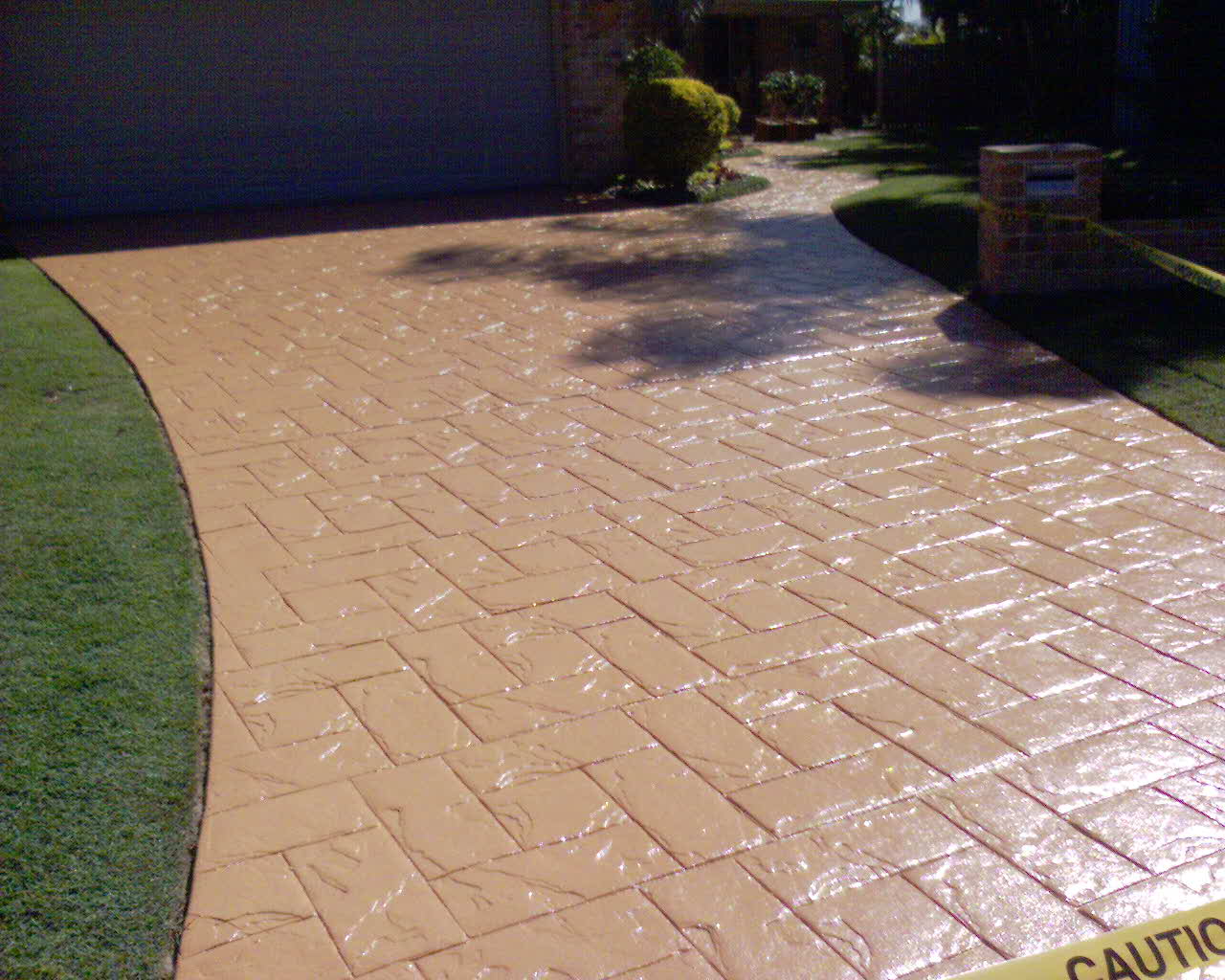 A glossy, tan-coloured stamped concrete driveway with a rectangular brick-like pattern curving toward a house.