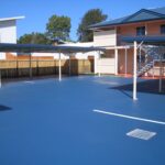 A bright blue paved parking lot with multiple metal-roofed carports and a two-storey beige apartment building in the background.