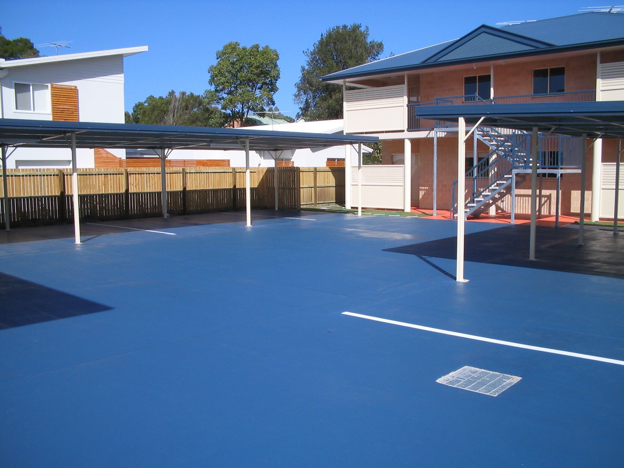 A bright blue paved parking lot with multiple metal-roofed carports and a two-storey beige apartment building in the background.