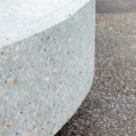 A curved, light-coloured polished concrete structure featuring visible stone aggregate, situated next to a textured pebble-dash ground surface.