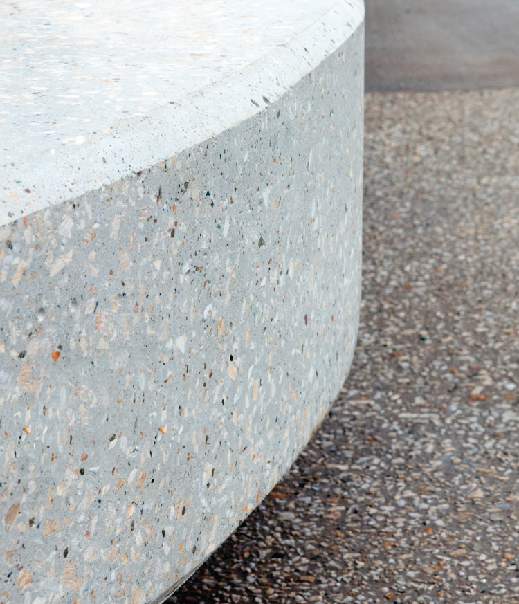 A curved, light-coloured polished concrete structure featuring visible stone aggregate, situated next to a textured pebble-dash ground surface.
