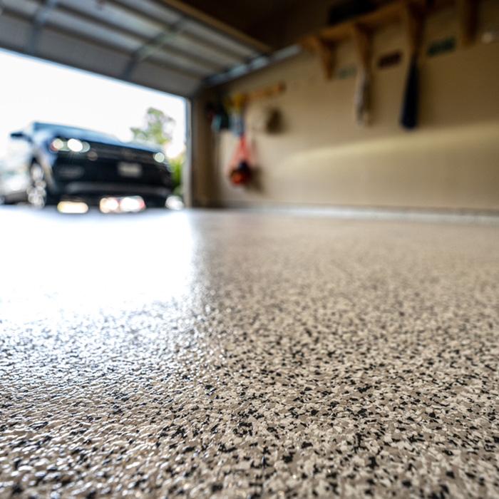 A low-angle, close-up view of a textured epoxy garage floor with tan, black, and white flakes, showing a blurred car and garage interior in the background.