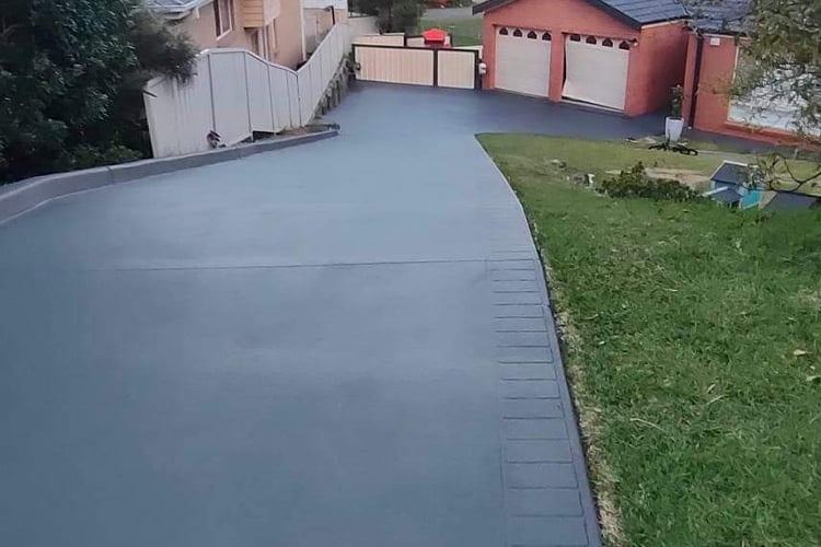 A smooth, dark charcoal grey resurfaced driveway sloping down toward a house, featuring a clean, uniform finish and a stamped border on the right edge.