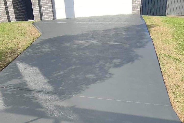 A smooth, grey concrete driveway leading to a garage, bordered by green grass and a brick house.