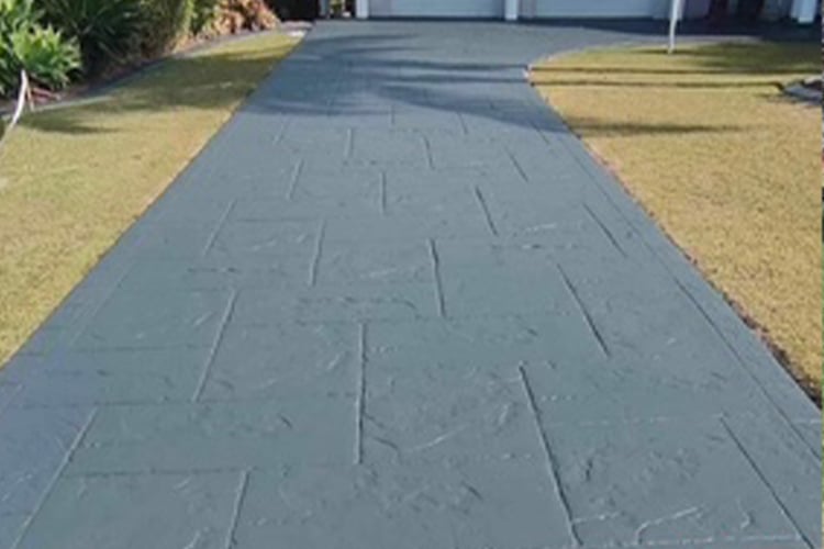A long, outdoor driveway made of stamped concrete with an ashlar slate pattern in a dark charcoal grey colour, bordered by grass.