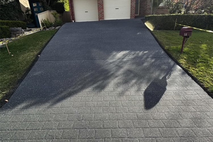 A dark grey, patterned driveway leads up to a brick house with two white garage doors, flanked by green lawns and a mailbox.