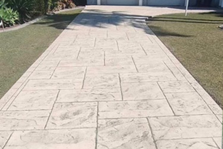 A long, outdoor driveway made of stamped concrete with an ashlar slate pattern in a light tan or beige colour, bordered by green grass.