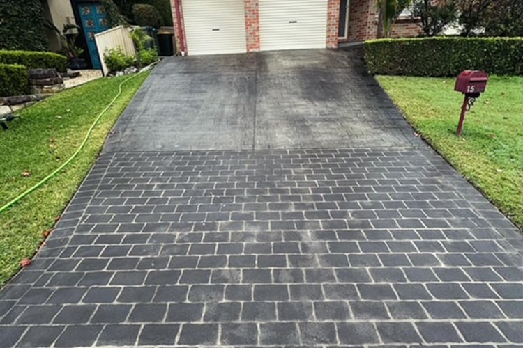 A worn, charcoal grey driveway showing significant fading and lighter patches on the upper section, leading to a brick house with two roller doors.