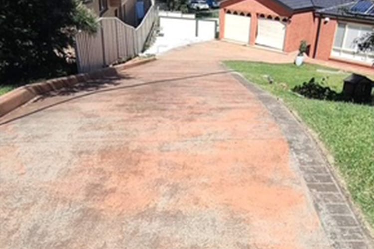 A steep, downward-sloping concrete driveway with a faded reddish-orange tint leading toward a brick house with white garage doors.
