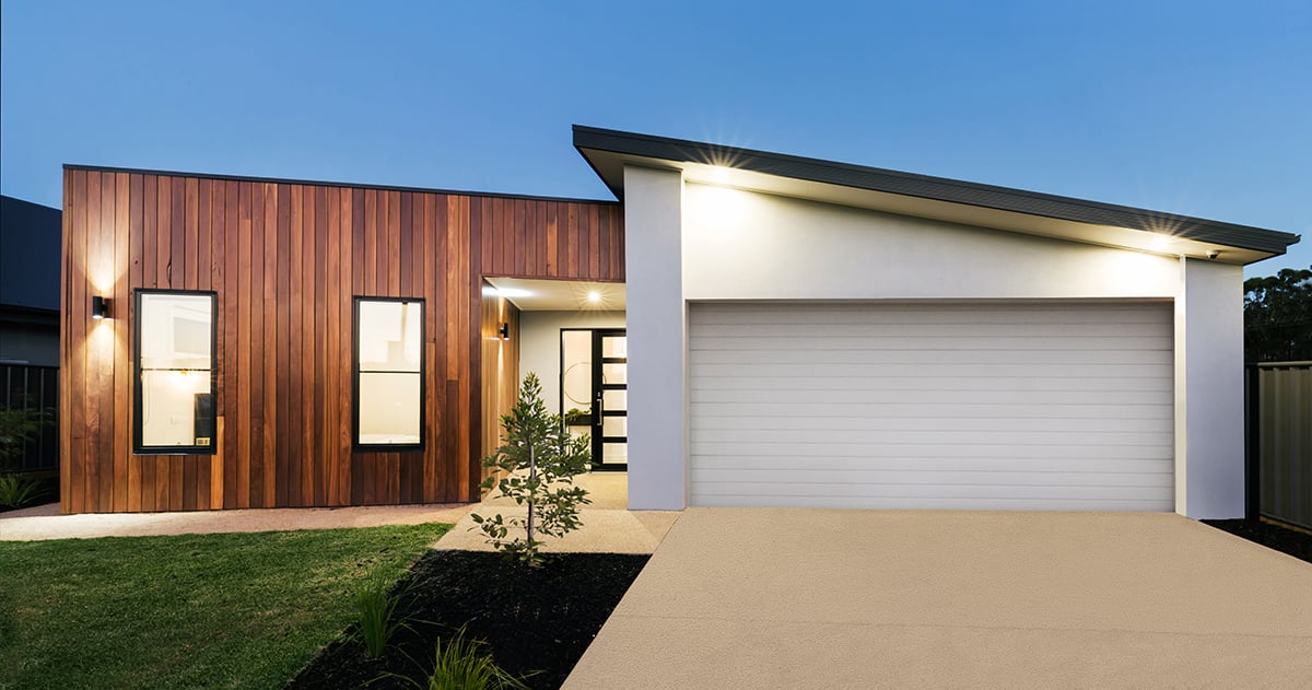 A modern, single-storey home at dusk featuring a facade of vertical wood panelling and white stucco, a wide tan driveway, and an integrated garage.