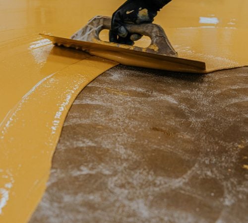A gloved hand using a notched trowel to spread a thick layer of bright yellow epoxy or resin coating onto a grey concrete floor.