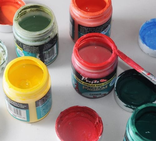 Several open jars of colourful acrylic paint, including yellow, red, and green, scattered on a white surface with a paintbrush.
