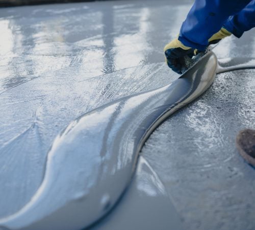 A person wearing blue workwear spread a thick, glossy grey epoxy coating over a concrete floor.