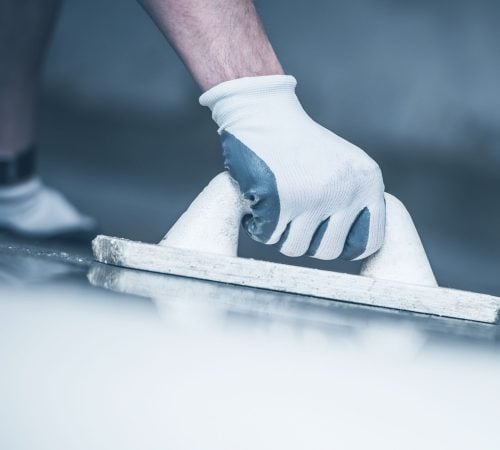A gloved hand using a white hand trowel to smooth a reflective, wet surface, likely during flooring or plastering work.