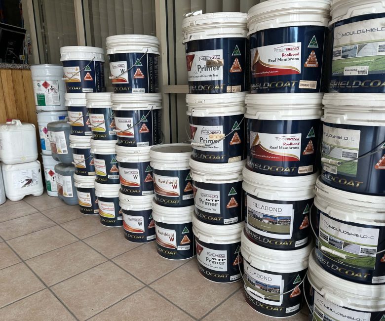 Stacks of large white and blue Shieldcoat buckets containing various roofing products like primer, roof membrane, and mould shield, arranged on a tiled floor.